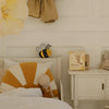 A cozy kids' room features a bed with a sunburst pillow, a doll, the Handmade Wooden Mini Lamp - Happy Bee as a whimsical night light, a gingham dress, the book "Who am I?", a bunny plush, and a white nightstand.