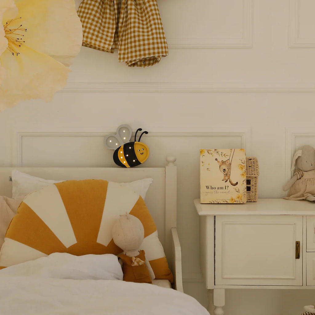 A cozy kids' room features a bed with a sunburst pillow, a doll, the Handmade Wooden Mini Lamp - Happy Bee as a whimsical night light, a gingham dress, the book "Who am I?", a bunny plush, and a white nightstand.