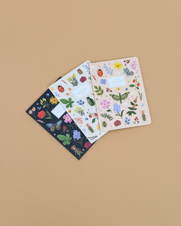 The Assorted Set of 3 Curio Notebooks, each with unique gold foil floral and botanical designs on the cover, is displayed in a fan arrangement on a beige background. Each ruled notebook showcases distinct colorful plant patterns.