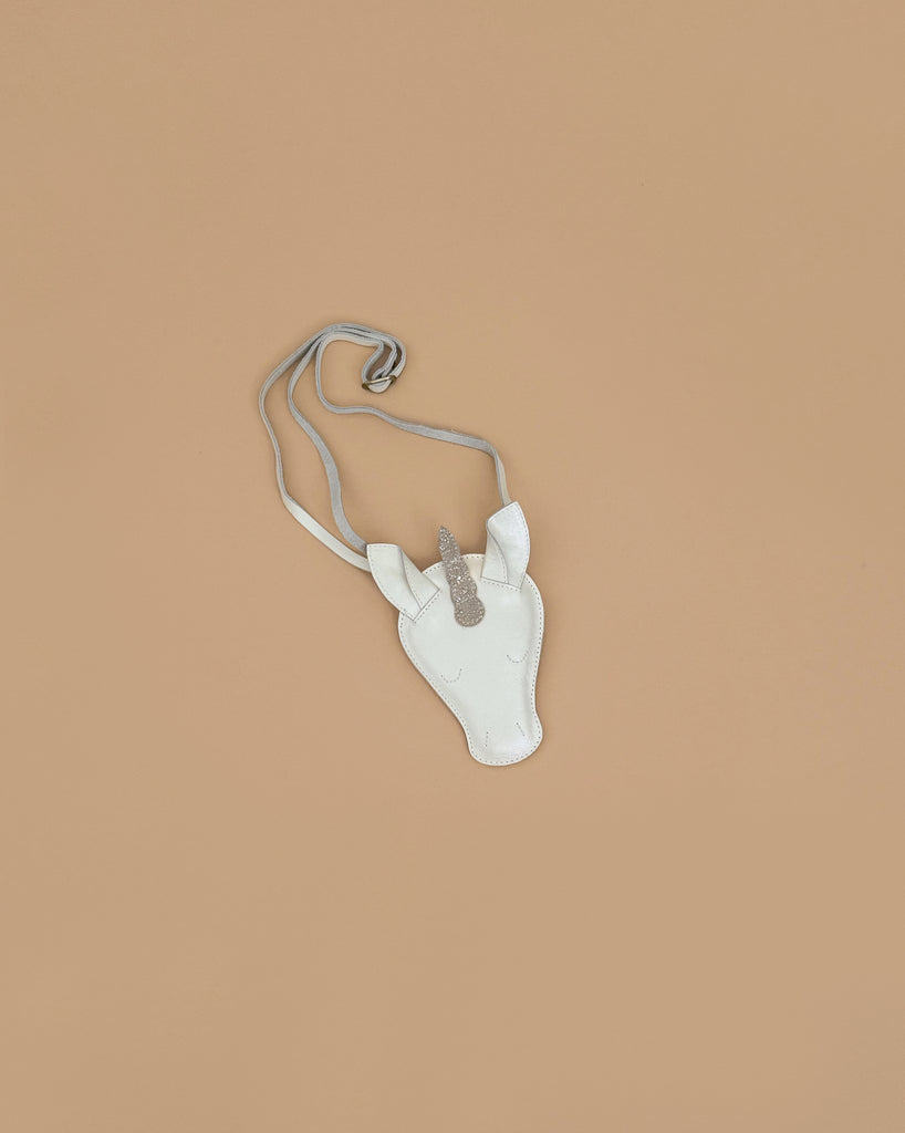 The Donsje Leather Blinc Purse - Unicorn is a small white purse with a silver glittery horn and adjustable leather strap, shown on a plain light brown background.
