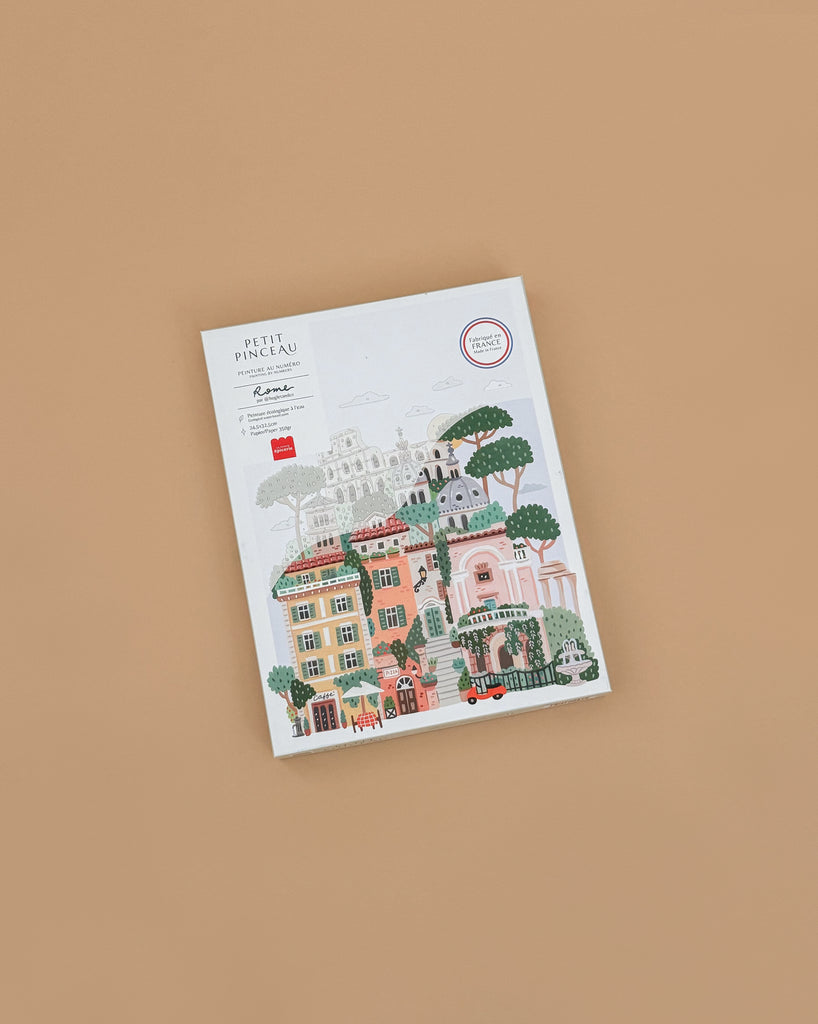 The "Paint By Number Kit - Rome" rests on a plain brown surface. Its vibrant cover features charming Roman buildings, trees, and a red scooter—perfect for aspiring artists seeking creative inspiration with a whimsical cityscape.