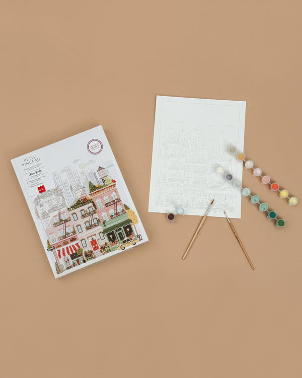 The Paint By Number Kit - Rome includes a colorful city scene on a light brown canvas, two paintbrushes, and paint pots—perfect for aspiring artists to create vibrant artwork.