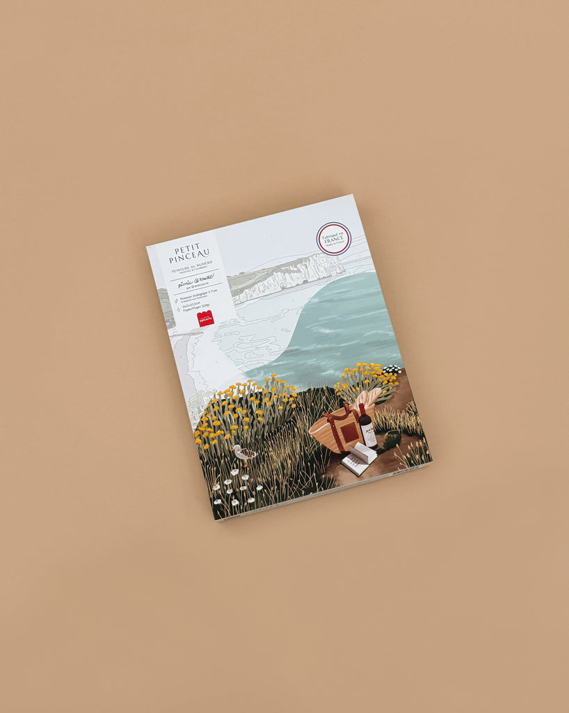 A paint by number kit, “Picnic in Étretat” by Ananya Bhandari, features an illustrated cover of the Normandy coast with wildflowers, a picnic basket, and books on a tan background, evoking the charm of La Petite Epicerie.