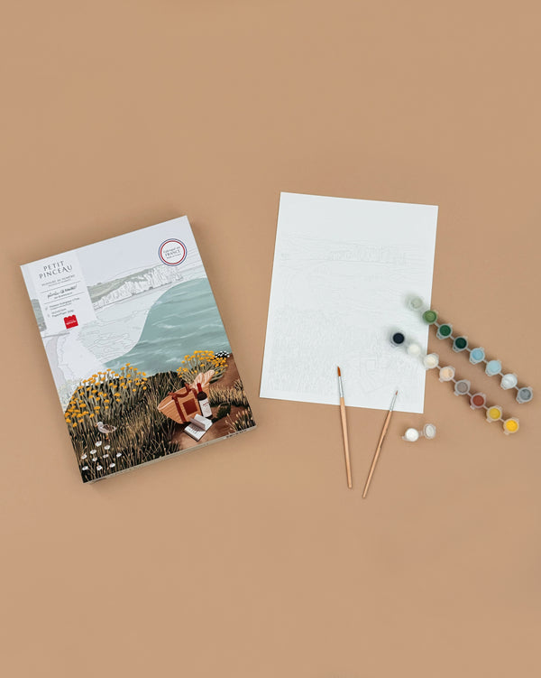 A flat lay of the Paint By Number Kit - Picnic in Étretat by Ananya Bhandari, shown with a paint set, two brushes, a pre-outlined blank canvas, and a reference image, all arranged on a light brown background.