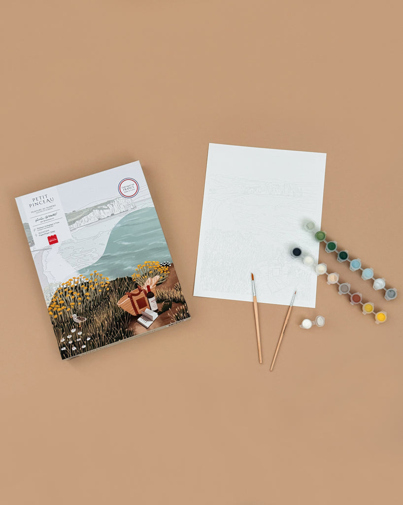 A flat lay of the Paint By Number Kit - Picnic in Étretat by Ananya Bhandari, shown with a paint set, two brushes, a pre-outlined blank canvas, and a reference image, all arranged on a light brown background.