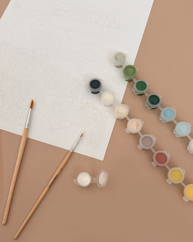 Two paintbrushes, small containers of paint, and the "Paint By Number Kit - Picnic in Étretat by Ananya Bhandari" are arranged on a beige surface, ready to inspire creativity with scenes from the Normandy coast.
