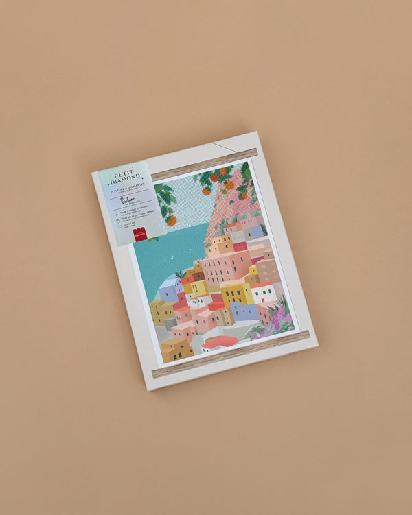 Framed colorful illustration of a coastal village with pastel houses, blue sea, and greenery, inspired by the Diamond Painting Kit - Positano by Petra Lidze, displayed against a plain beige background.