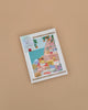 Framed colorful illustration of a coastal village with pastel houses, blue sea, and greenery, inspired by the Diamond Painting Kit - Positano by Petra Lidze, displayed against a plain beige background.