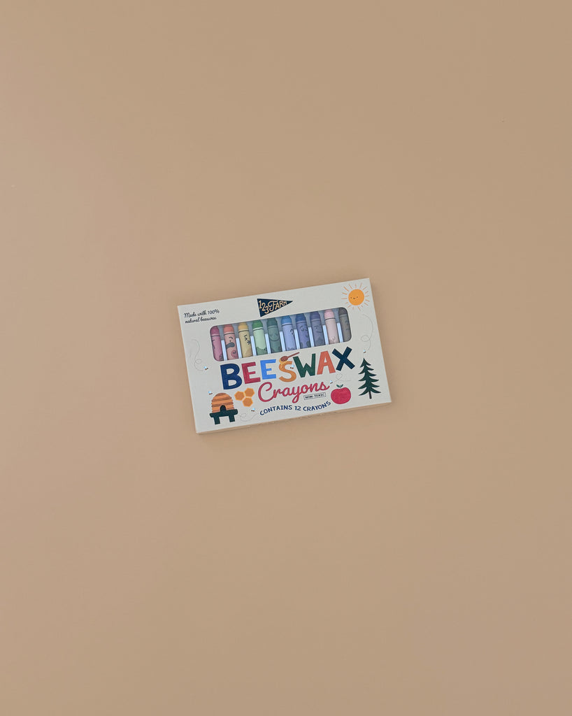 A box of 12 Beeswax Crayons – Non-Toxic with colorful sticks visible through playful, brightly illustrated packaging sits on a plain beige surface.