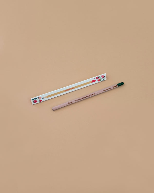 A Strawberry Plantable Pencil – Eco-Friendly with a green tip rests beside a white box adorned with red strawberries and leaves, both crafted from sustainable materials, set on a light brown background.