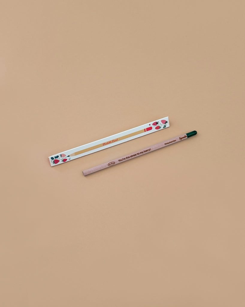 A Strawberry Plantable Pencil – Eco-Friendly with a green tip rests beside a white box adorned with red strawberries and leaves, both crafted from sustainable materials, set on a light brown background.