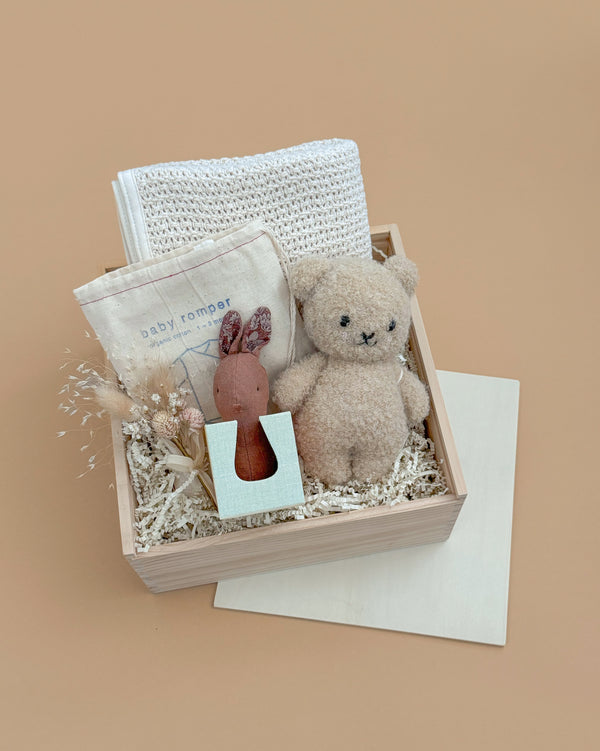 The Newborn Gift Set features an organic cotton baby romper, plush teddy bear, bunny rattle, white cloth, and dried flowers in a wooden box with shredded paper on a beige background.