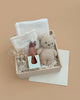 The Newborn Gift Set features an organic cotton baby romper, plush teddy bear, bunny rattle, white cloth, and dried flowers in a wooden box with shredded paper on a beige background.