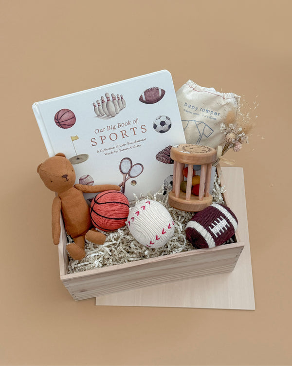 The Newborn Gift Set includes a sports-themed children's book, a brown Maileg Teddy, basketball, baseball and football toys, a handmade wooden rattle, dried flowers, and a folded baby blanket on a beige background.