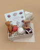The Newborn Gift Set includes a sports-themed children's book, a brown Maileg Teddy, basketball, baseball and football toys, a handmade wooden rattle, dried flowers, and a folded baby blanket on a beige background.
