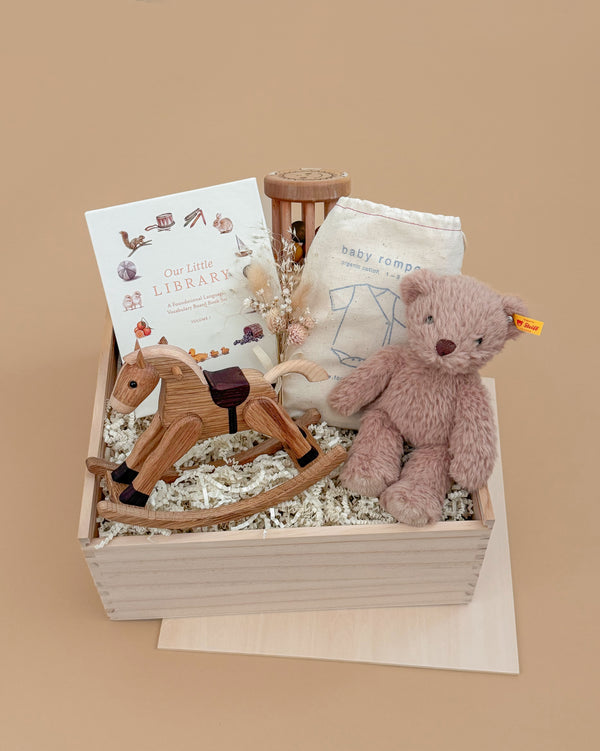 The Newborn Gift Set features a wooden box with a plush teddy bear, rocking horse, organic cotton romper in a cloth bag, children's book, and dried flowers on shredded paper, set against a soft beige background.