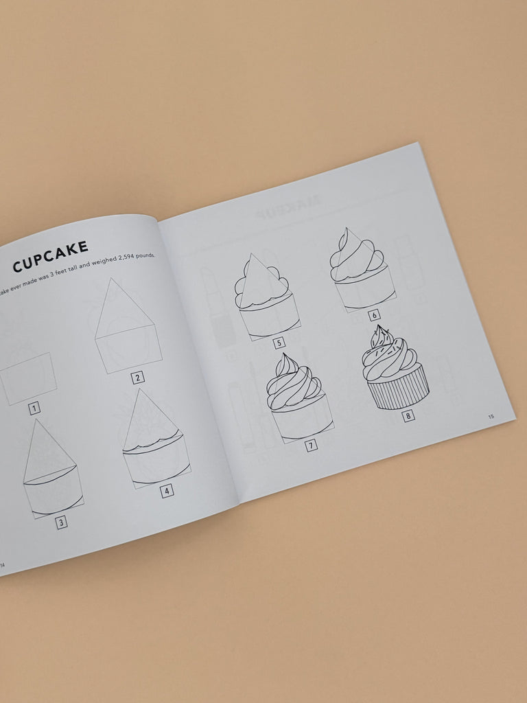 An open copy of "All the Things: How To Draw Books For Kids" rests on a beige surface, displaying step-by-step cupcake drawing instructions, from basic shapes to a finished treat—ideal for simple drawing lessons.