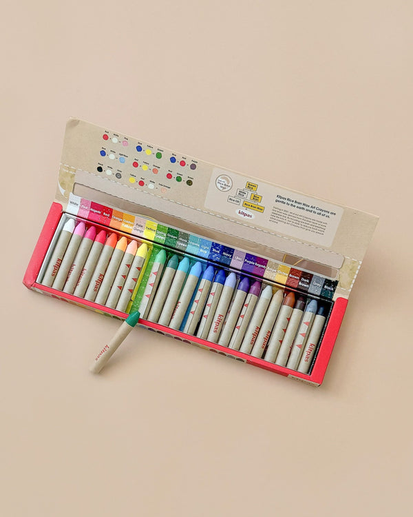 An open box of Kitpas Crayons - 24 Colors, made in Japan, is displayed on a beige surface with the crayons neatly arranged and a single green crayon lying diagonally across the set.