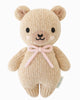 The Cuddle + Kind Baby Honey Bear - Blush is a beige knitted teddy, handmade in Peru from Peruvian cotton yarn, featuring black eyes, a small nose and mouth, pink blush cheeks, and a pink yarn bow—shown against a plain white background.