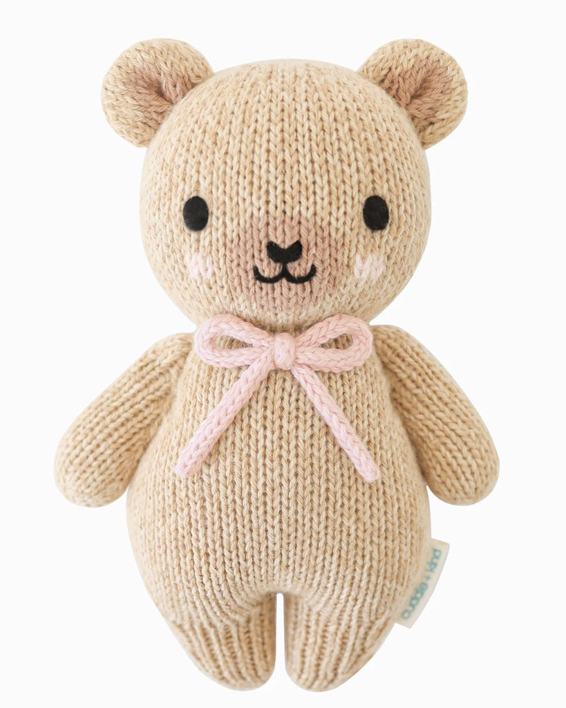The Cuddle + Kind Baby Honey Bear - Blush is a beige knitted teddy, handmade in Peru from Peruvian cotton yarn, featuring black eyes, a small nose and mouth, pink blush cheeks, and a pink yarn bow—shown against a plain white background.