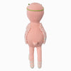 Cuddle + Kind Penelope the Flamingo is a pink, hand-knit doll with a round head, small tail, green headband, fluffy vest, and white crisscross ribbons on its feet. The doll stands upright facing away.