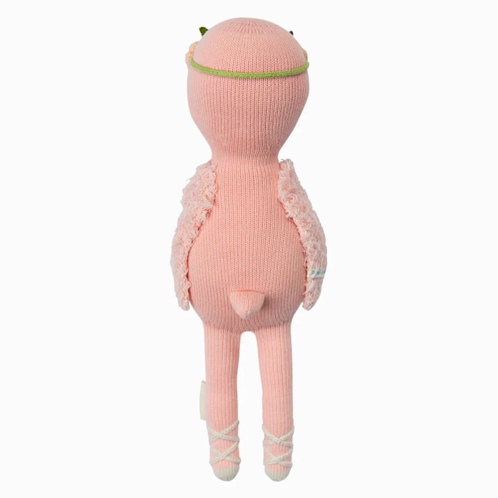 Cuddle + Kind Penelope the Flamingo is a pink, hand-knit doll with a round head, small tail, green headband, fluffy vest, and white crisscross ribbons on its feet. The doll stands upright facing away.