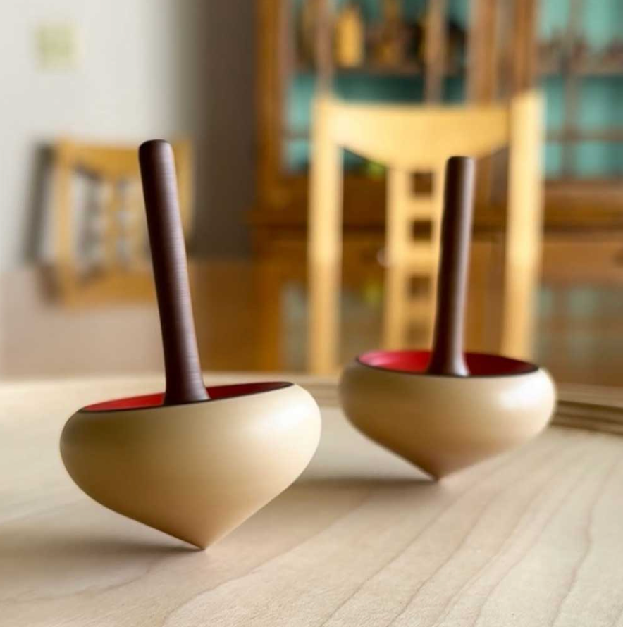Two Mader Zwirbel Spinning Tops - Red, featuring dark handles and light, rounded bodies crafted from Walnut and Ash, stand upright on a smooth, light wooden surface. A wooden chair and cabinet appear blurred in the background.