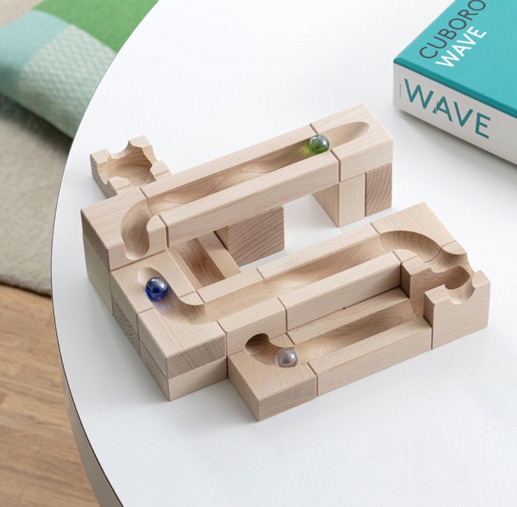 The Cuboro Wave Marble Run Extra Set is shown assembled on a white table with three marbles, interconnected tracks, its blue-and-white "WAVE" box, and part of a green blanket visible nearby.