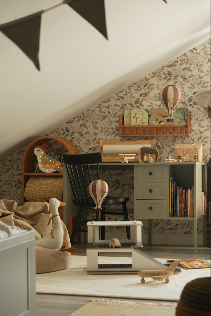 A cozy attic children's room with floral wallpaper, a green desk full of books, a black chair, stuffed toys, a Hot Air Balloon - Light Brown / Dark Brown hanging from the ceiling, and a soft beige bean bag under the sloped roof.