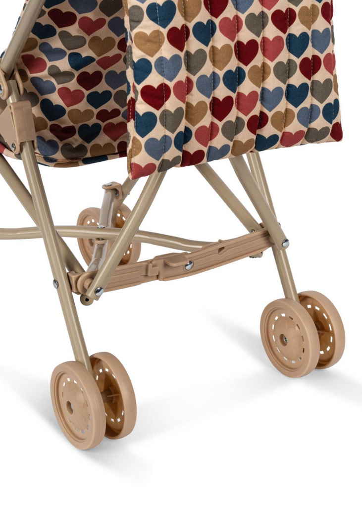 A close-up of the Konges Sloejd Doll Stroller - Heart Kisses, featuring tan double wheels, a sturdy frame, and a fabric canopy adorned with rows of blue, red, and tan hearts.