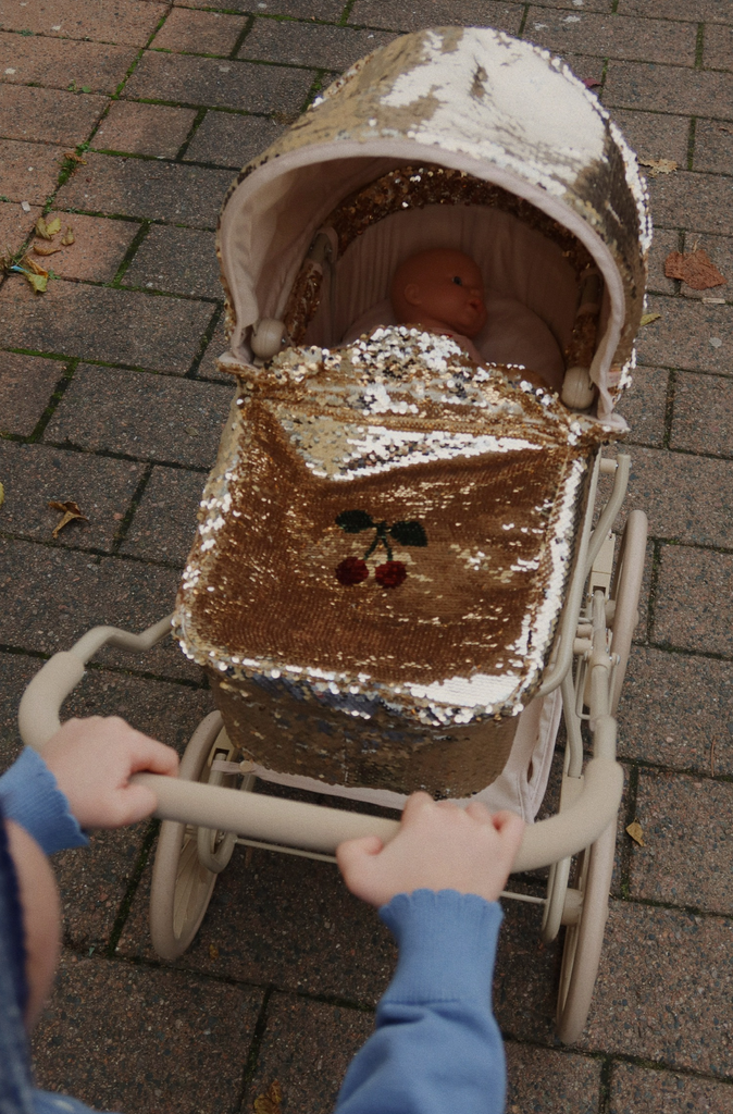 A child pushes the Konges Sloejd Sparkly Doll Pram - Golden Cherry, featuring a gold sequin cover with cherry design, while carrying a doll outdoors on a paved walkway.