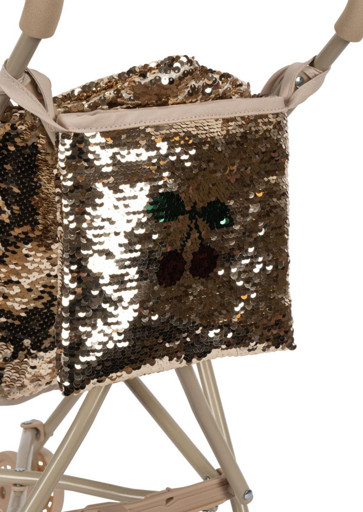 A close-up of the Konges Sloejd Sparkly Sequin Doll Stroller - Golden Cherry, featuring a shiny gold sequin bag with red cherry and green leaf design attached to a beige sparkly stroller.