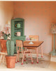 The Maileg Fully Furnished Farmhouse dining room includes a small wooden table with desserts, two chairs, potted cactus, green cabinet with cups, patterned rug, and pink patterned wallpaper for a charming touch.