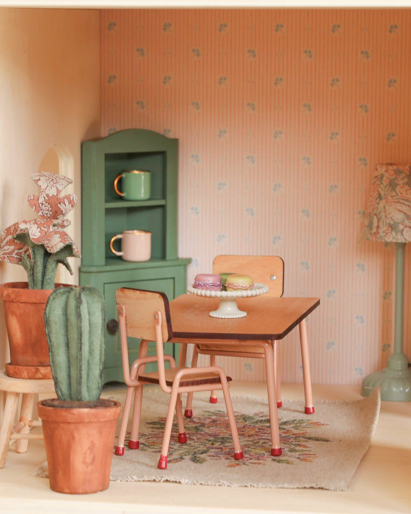 The Maileg Fully Furnished Farmhouse dining room includes a small wooden table with desserts, two chairs, potted cactus, green cabinet with cups, patterned rug, and pink patterned wallpaper for a charming touch.