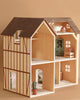 The Maileg Fully Furnished Farmhouse features a dark brown roof, beige walls with brown trim, multiple rooms filled with miniature furniture, and a small potted cactus by the front door, all set against a light brown background.