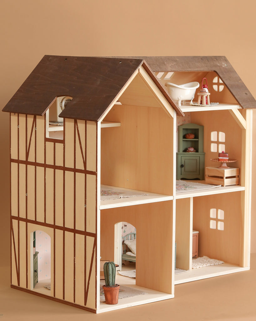The Maileg Fully Furnished Farmhouse features a dark brown roof, beige walls with brown trim, multiple rooms filled with miniature furniture, and a small potted cactus by the front door, all set against a light brown background.