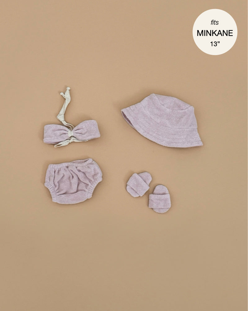 A flat lay of the Minikane Doll Clothing | Emile & Ida Ursula 3-piece Swimsuit in Petal terrycloth, featuring a knotted top, bloomers, and bucket hat for 13" Minikane dolls, displayed on a tan background.