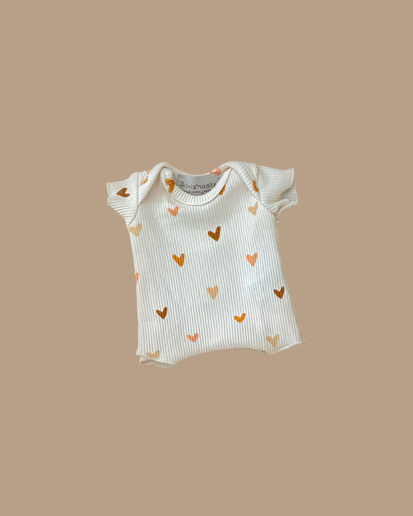 The Minikane Doll Clothing Les P'tits Basique Ribbed Bodysuit in Hearts is a beige, cream-colored short-sleeve onesie made from soft cotton with golden and brown heart patterns for Minikane 13" & 14" dolls. Designed in France, it fits Gordis dolls.