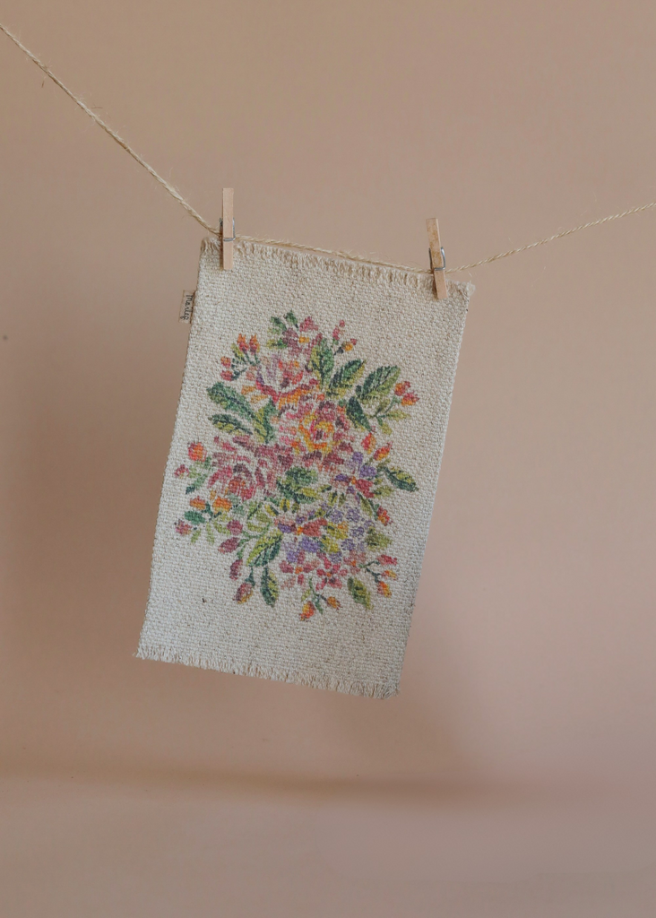 A piece of fabric with a colorful embroidered floral design hangs from a clothesline by two wooden clothespins, set against a peach background, softly illuminated by the Maileg Living Room Set.