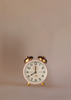 A small vintage white alarm clock with gold bells and legs sits beside the Maileg Dancing Mouse in Daybed – Cozy Morning Set, both arranged on a plain beige background with the clock displaying 12:00.