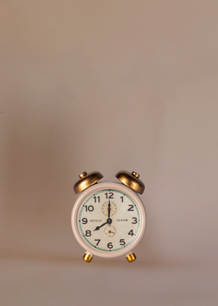 A small vintage white alarm clock with gold bells and legs sits beside the Maileg Dancing Mouse in Daybed – Cozy Morning Set, both arranged on a plain beige background with the clock displaying 12:00.