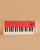 The Loog Piano is a compact, minimalist digital piano featuring wooden sides, black and white velocity-sensitive keys, a bright red top panel, and a single large knob on the right, shown against a plain beige background.