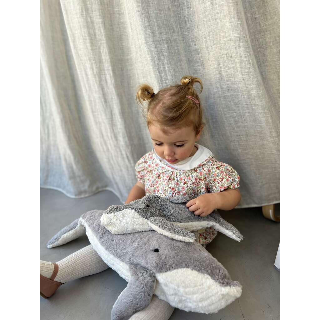 A young child with light brown hair in two pigtails sits on the floor, wearing a floral dress and holding a Senger Naturwelt Cuddly Animal - Whale made from organic cotton. A light curtain hangs in the background.
