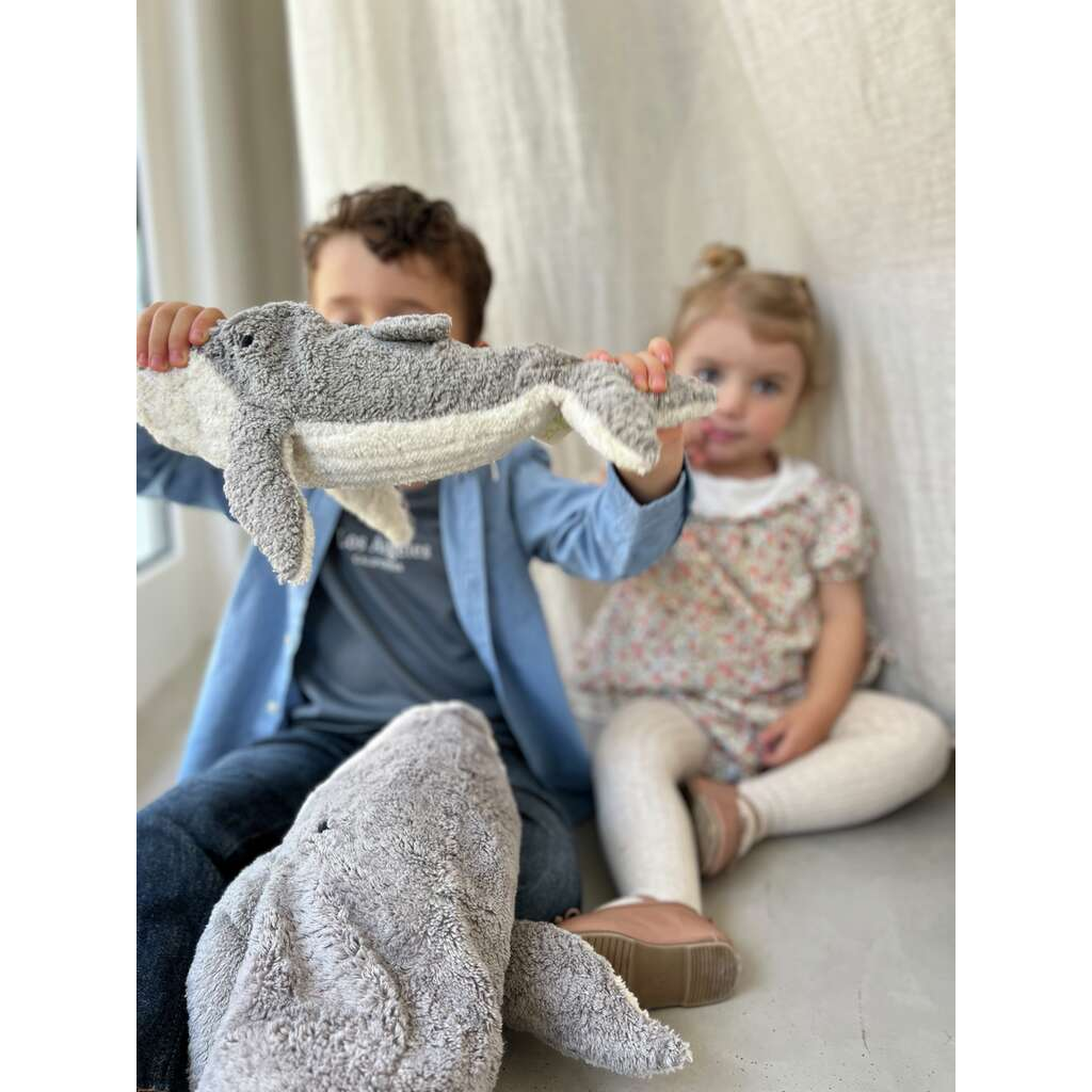 Two young children sit indoors by a window. The boy in front holds up a Senger Naturwelt Cuddly Animal - Whale, partially covering his face, while the girl behind him looks at the camera. Another plush whale rests on the floor nearby.