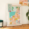 The Diamond Painting Kit - Positano by Petra Lidze showcases a vibrant seaside hillside with stacked houses, shown on a wooden surface beside a green candle and leafy plant.