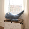 The Ride-On Whale Pouf, a large and soft gray seat with a playful whale design and rope loop, sits atop a white wooden chest by a window with sheer curtains, letting in natural light.