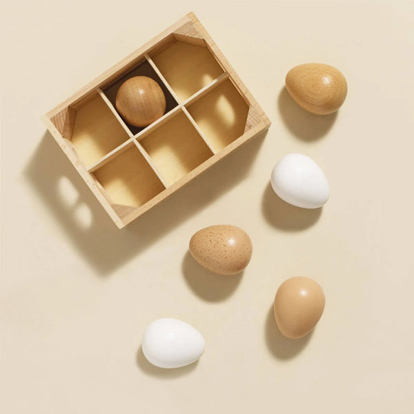 The Farm Eggs Wooden Market Crate includes a wooden box with six compartments holding one wooden egg. Five extra wooden toy eggs, in white and natural wood tones, are displayed nearby for a plastic-free play food collection.