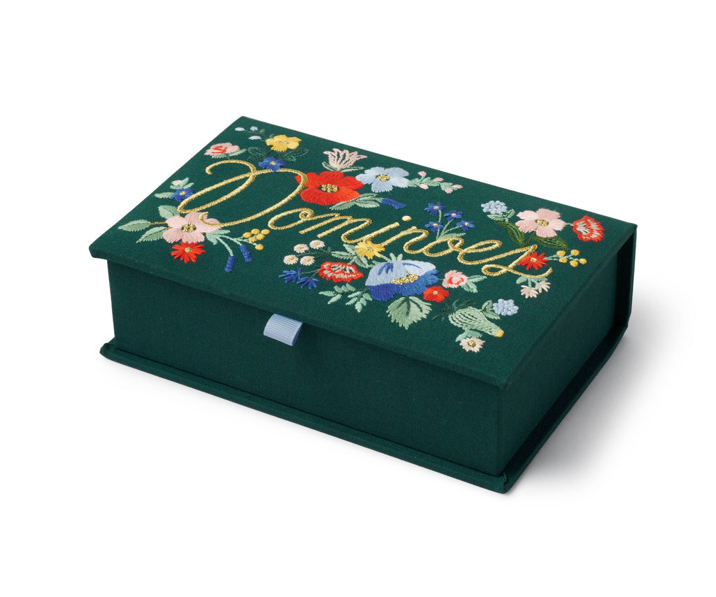 The Strawberry Fields Dominoes Set is a dark green embroidered keepsake box featuring colorful illustrations and "Dominoes" stitched in yellow cursive on the lid, with a light blue ribbon tab on the front.