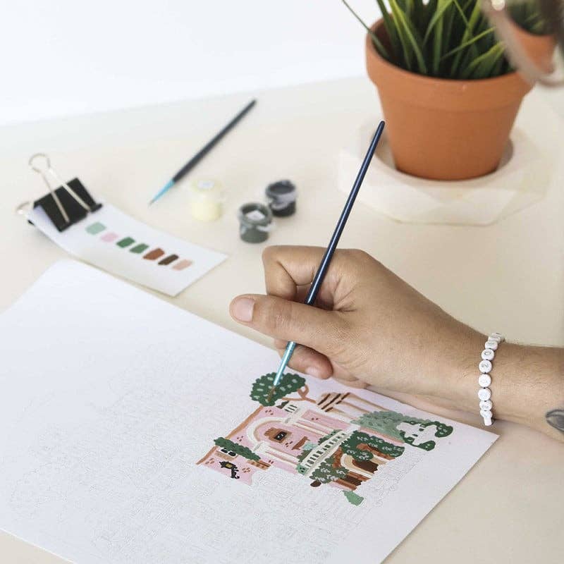 An aspiring artist paints a vibrant Rome scene from the Paint By Number Kit – Rome, using a thin brush. On the table: paint pots, a color swatch card, and a potted plant. The artist sports a white beaded bracelet.