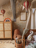A cozy children’s bedroom with floral wallpaper, the Hot Air Balloon - Burgundy wooden decor, a rattan wardrobe, a wicker basket, rainbow light, bunting, and a wooden bed with plush toys and cushions—perfect for charming children's room decoration.